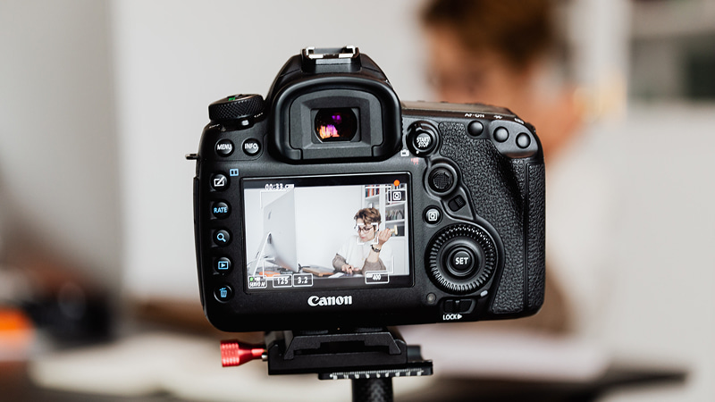 ビデオ収録中のキヤノン（Canon）製カメラ。モニターには被写体の女性と設定値が映り、本格的なコンテンツ制作の裏側を演出。