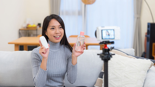 リビングのソファに座り、カメラに向かって化粧品のボトルを紹介している笑顔の女性。三脚に固定されたカメラで動画撮影中であり、Vlog制作やライブコマースの様子を伝えています。
