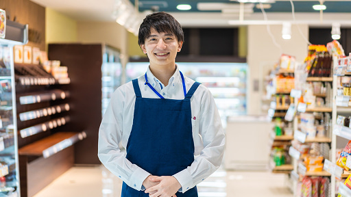 コンビニエンスストアの店内で、青いエプロンを着用して笑顔で立つ男性店員。背景には整然と並んだ商品棚があり、清潔感のある接客イメージや店舗スタッフの紹介に適しています。
