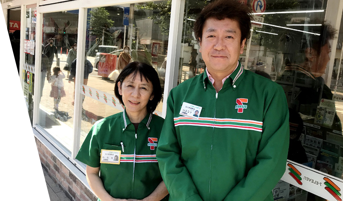 セブン-イレブンの緑色のユニフォームを着用し名札をつけた、笑顔で並ぶ男女のスタッフ。日差しの差し込む明るい店舗のガラス窓を背景に、親しみやすさと清潔感のある雰囲気でこちらを見つめて立っている様子。