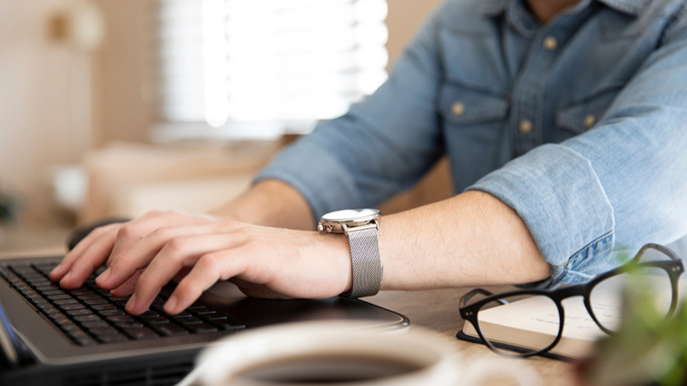 デニムシャツを着た人物が、デスクの上でノートPCのキーボードをタイピングしている手元のクローズアップ。横には眼鏡とノート、手前にはコーヒーカップが置かれた落ち着いたワークスペースの風景。