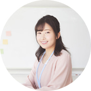 ホワイトボードの前で、ピンクのブラウスを着て穏やかに微笑む日本人女性講師